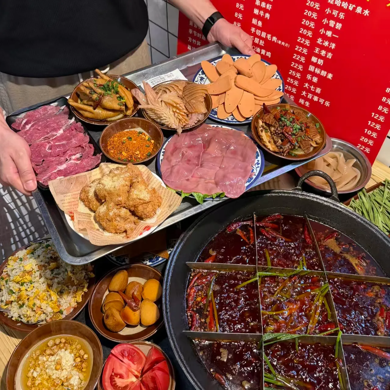 A hotpot table with a tray of raw plates, a bowl of sesame-oil dip, fried pork and egg fried rice, and a price board on the wall behind