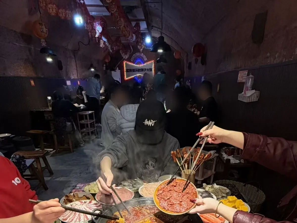Diners at tables inside a tunnel hotpot restaurant, under an arched rock ceiling hung with red banners