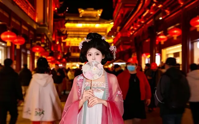 A woman in pink and green hanfu holding a round silk fan on a red-lantern-lit street at night in Shanghai, crowds walking behind her