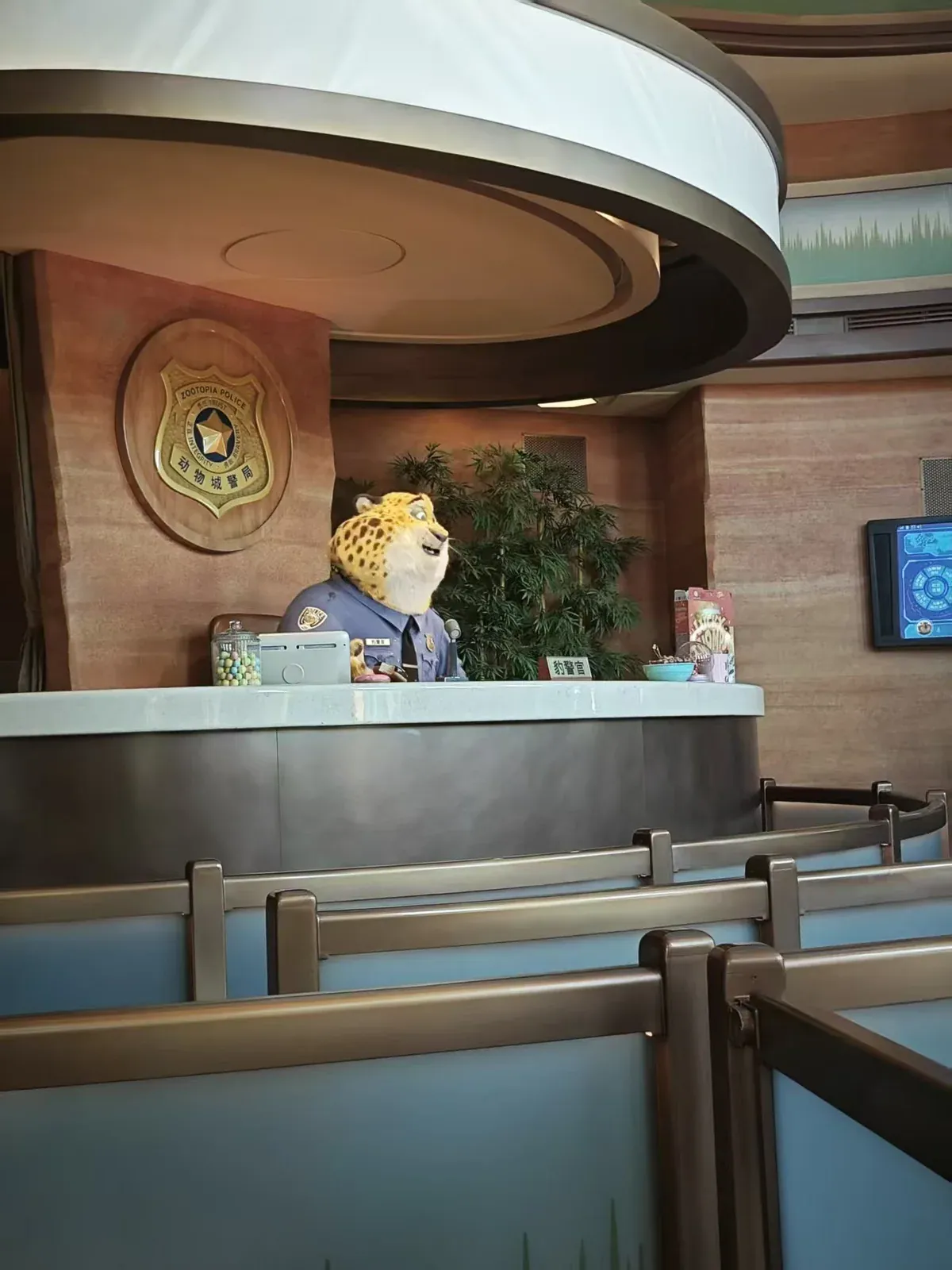 Officer Clawhauser, the cheetah receptionist, sitting behind the front desk at the Zootopia police station with silver queue barriers in the foreground